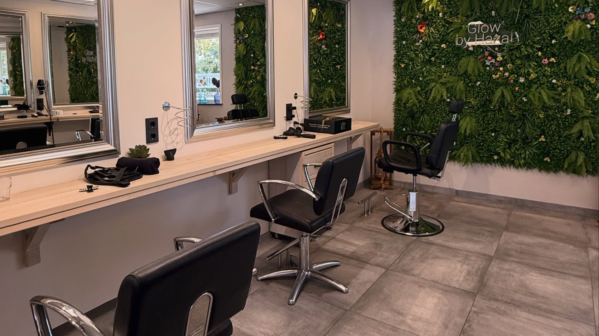 De salon van binnen: spiegels, zwarte stoelen, een houten werkblad en de groene bloemenwand